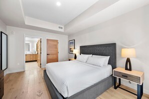 A modern bedroom with a large bed, wooden flooring, a nightstand with a lamp, and a door leading to a bathroom.