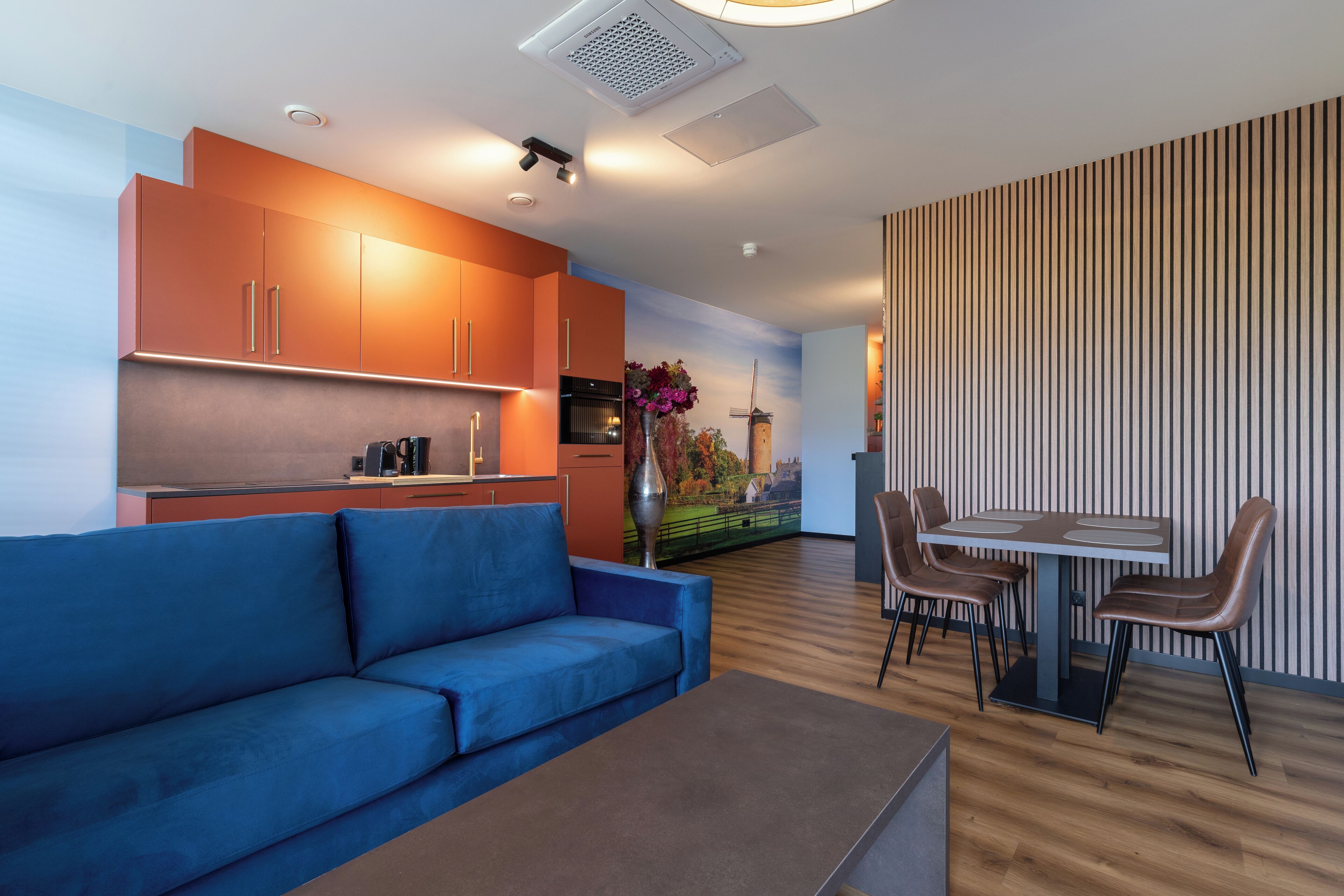 Un soggiorno moderno con un divano blu, un tavolo da pranzo con sedie e una zona cucina con mobili arancioni.