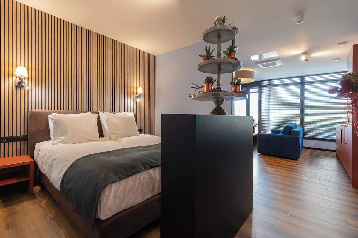 A modern hotel room with a large bed, a wooden accent wall, a built-in TV, and a large window with blinds.