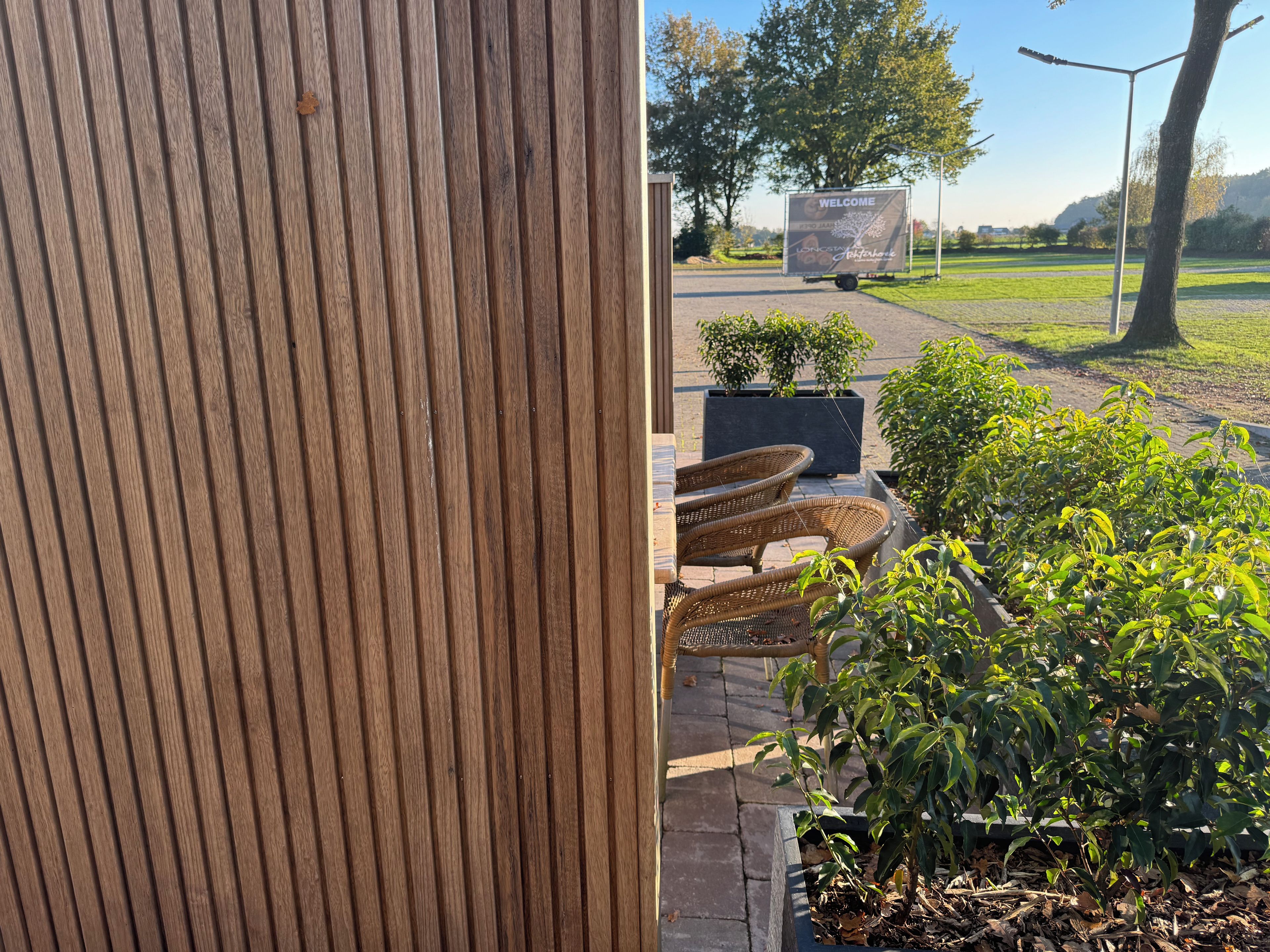 Eine Wand aus Holzverkleidung, ein Sitzbereich im Freien mit Korbstühlen und ein Willkommen-Schild in einem angelegten Garten.