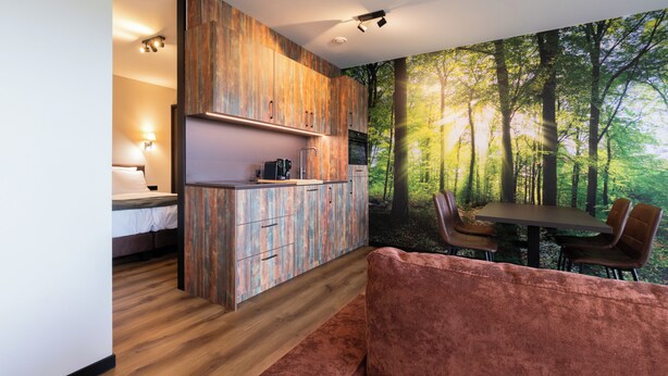 A modern kitchen with wooden cabinets, a dining area with a table and chairs, and a large forest mural on the wall. - Longstay Achterhoek (Zeddam)
