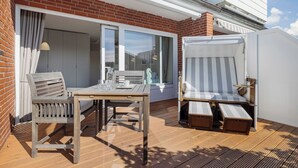 A patio with a wooden table and chairs, a striped lounge chair, and a sliding glass door leading to a balcony. - Pure island love! You can't book much more Sylt style (Wenningstedt-Braderup (Sylt))