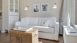 A white sofa with cushions, a wicker basket, a cabinet with glass doors, and two framed pictures on the wall. - Pure island love! You can't book much more Sylt style (Wenningstedt-Braderup (Sylt))