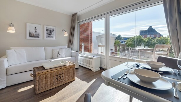 A modern living room with a white sofa, a dining table set with bowls and cutlery, and a large window with a view of a brick building and trees. - Pure island love! You can't book much more Sylt style (Wenningstedt-Braderup (Sylt))