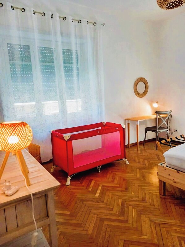 A room with a wooden herringbone floor, a red crib, a small table with a lamp, a bed, and a window with sheer curtains. - MAMACRI Holiday in Rome - for large groups (Roma)