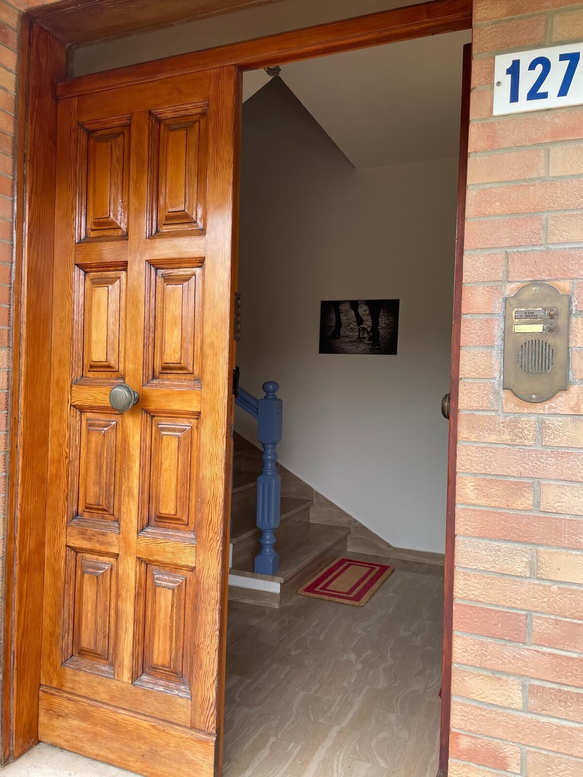 Una puerta doble de madera con picaporte de latón, un muro de ladrillo con buzón y una escalera que conduce a un piso superior.