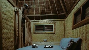 Ein kleiner, rustikaler Raum mit Holzboden und -wänden, einem Fenster und einem Bett mit blauem Kissen und zwei zusammengerollten Gegenständen darauf.