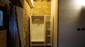 Une petite pièce étroite avec un mur en pierre, une porte en bois et une armoire blanche où sont suspendus des vêtements.