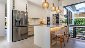 Een moderne keuken met een roestvrijstalen koelkast, een centraal kookeiland met barkrukken, hanglampen en uitzicht op een tuin door grote ramen.