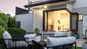 Een terras met een tafel en stoelen, een plant in pot, en een uitzicht op het interieur door een glazen deur.