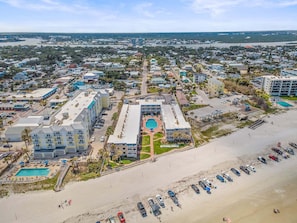 Um hotel à beira-mar com vários edifícios, uma piscina e uma área de praia com carros estacionados.