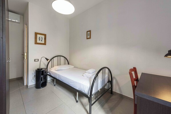 Una habitación pequeña y limpia con una cama individual, un escritorio y una silla. Hay un cubo de basura y un cuadro en la pared.