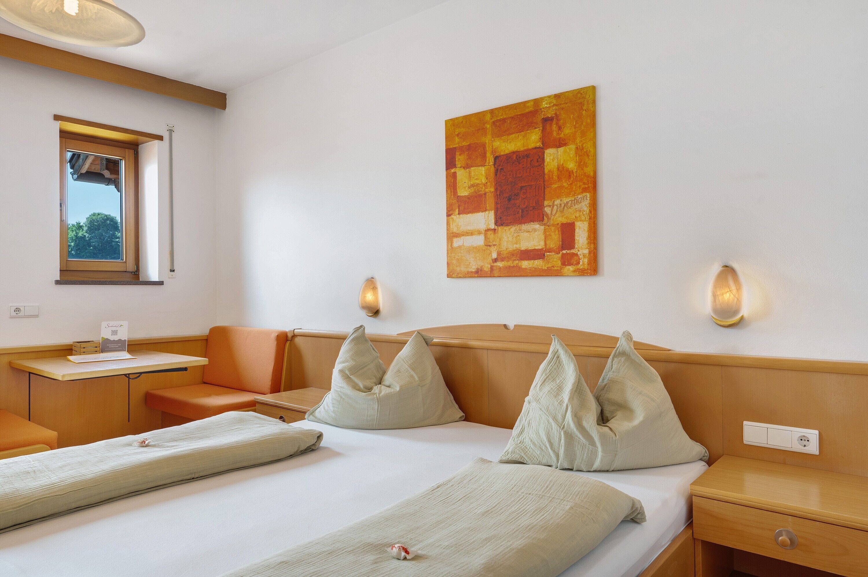 A hotel room with a bed, two pillows, a bedside table, a nightstand, a window, and a wall-mounted artwork.