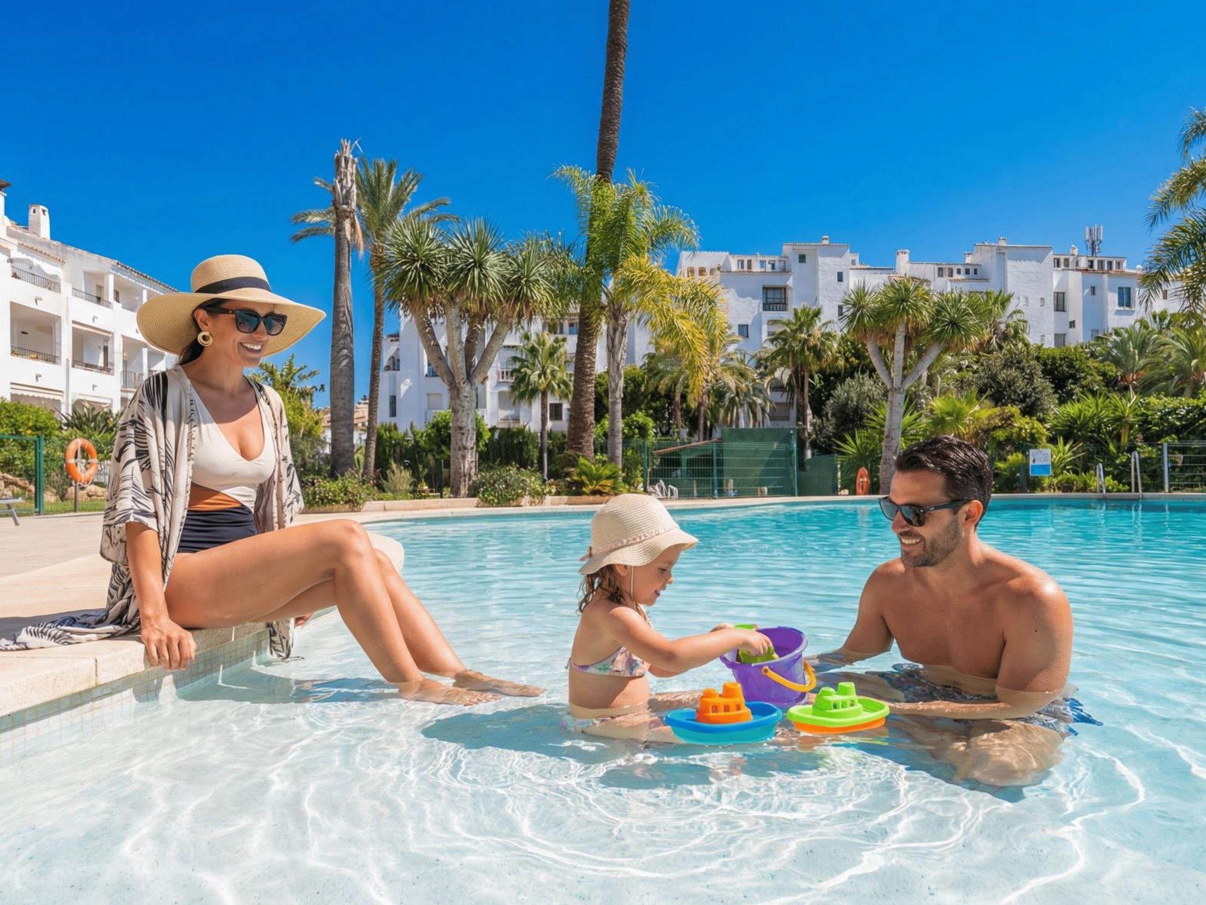Eine Familie am Pool, mit blauem Himmel, Palmen und weißen Gebäuden im Hintergrund.