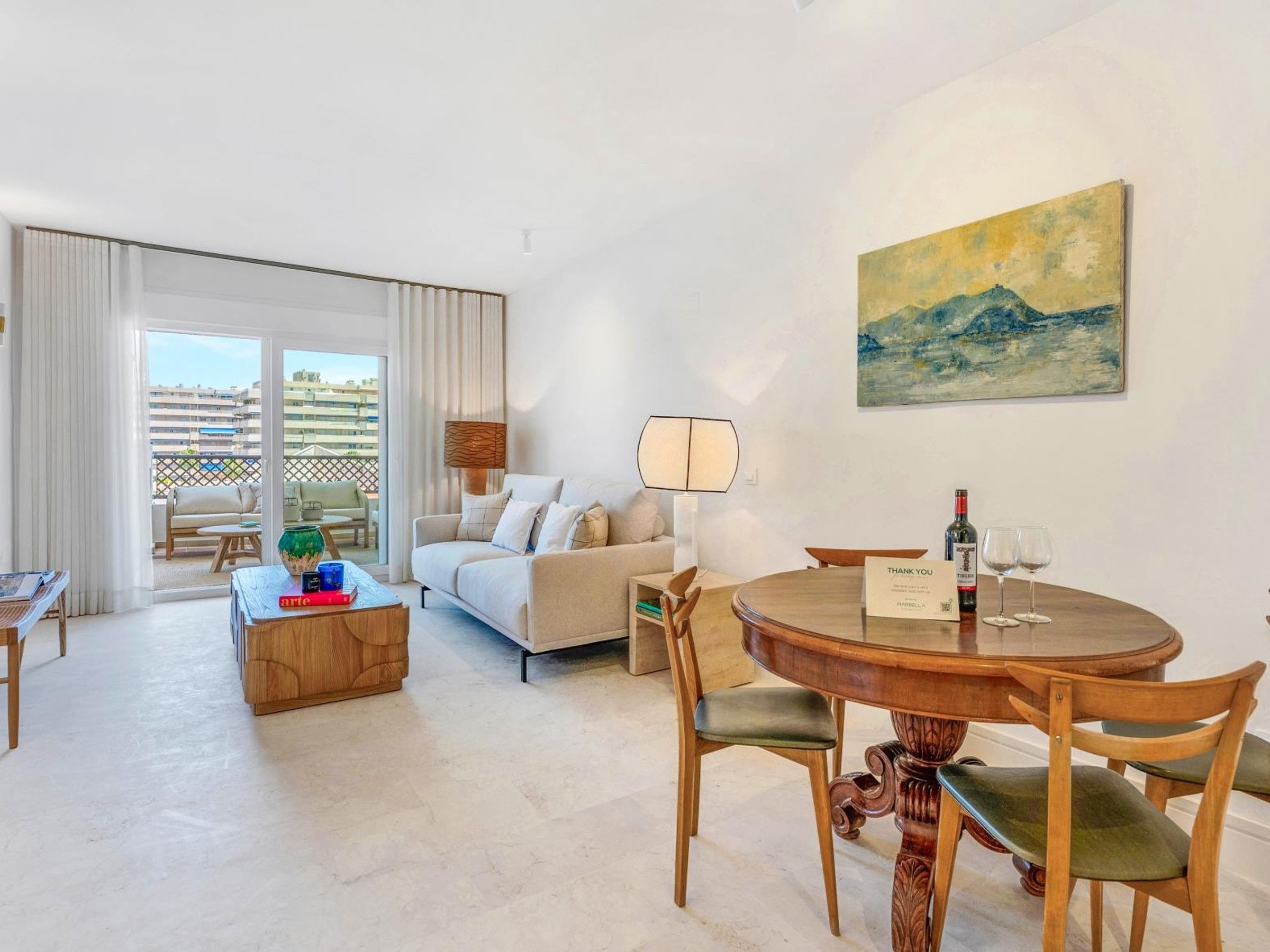 Ein modernes Wohnzimmer mit Sofa, Couchtisch und Esstisch. Ein Gemälde hängt an der Wand und vom Balkon aus ist ein Blick auf Gebäude möglich.