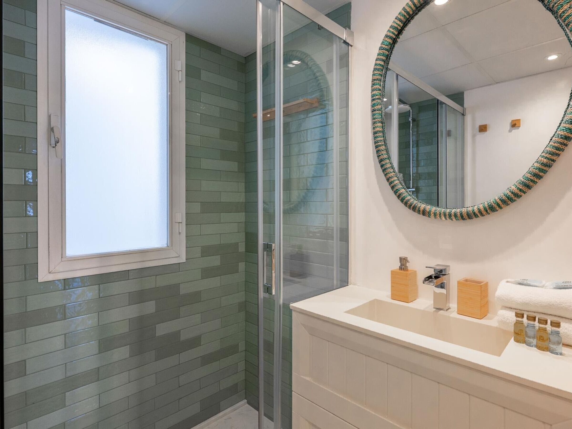 Un baño con cabina de ducha de vidrio, una ventana, un espejo con marco decorativo y tocador con artículos de aseo.