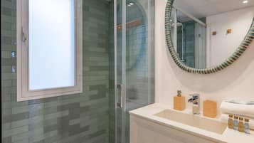 Un baño con cabina de ducha de vidrio, una ventana, un espejo con marco decorativo y tocador con artículos de aseo.