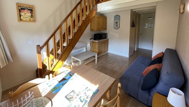 A spacious living area with a wooden staircase, a dining table with a placemat, a sofa, and a kitchen area in the background.