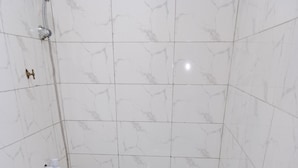 Une petite salle de bain blanche, équipée de toilettes, d’une douche et d’un lavabo. On y trouve deux seaux et une bouteille de savon liquide sur le sol.