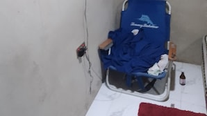 Une petite pièce avec un lit simple, une chaise recouverte d’une housse bleue, un téléviseur fixé au mur et un tapis rouge.