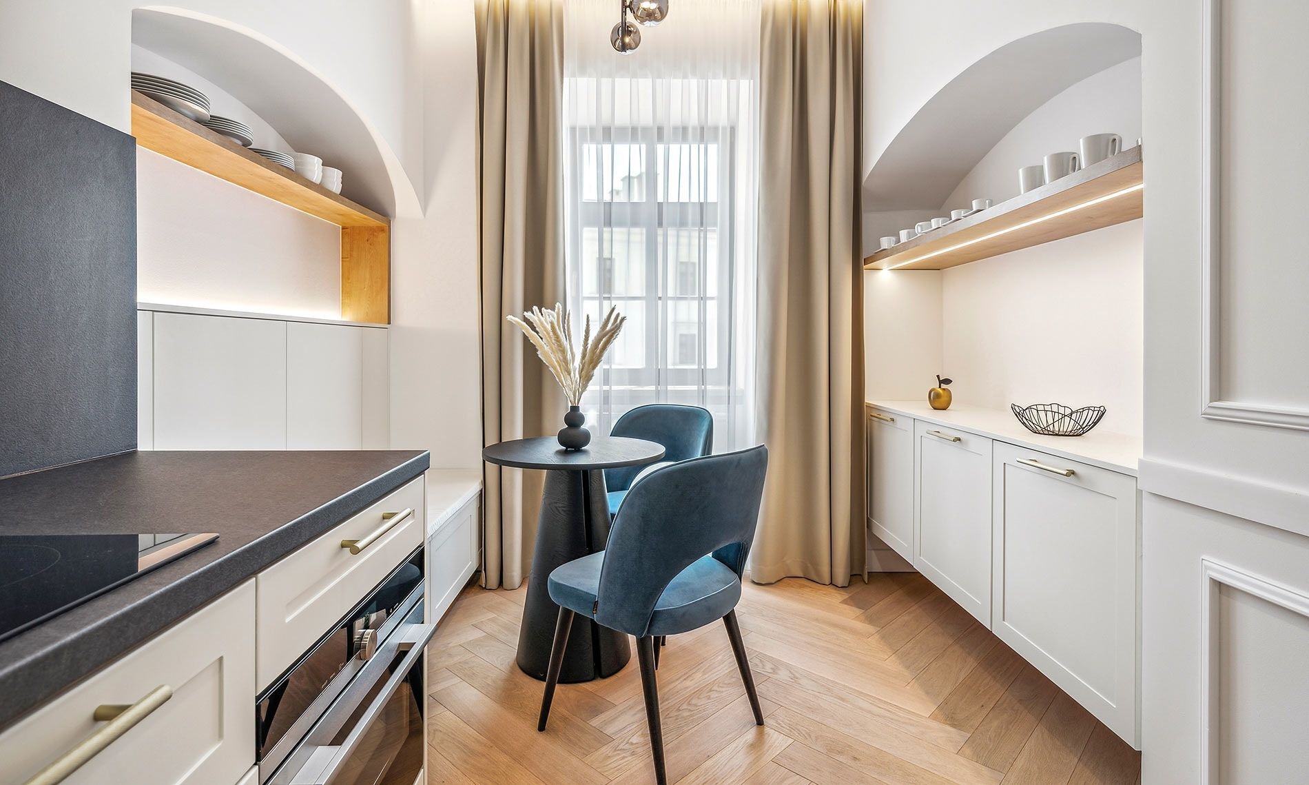Eine moderne Küche mit weißen Schränken, einer schwarzen Arbeitsplatte und einem runden Tisch mit Stühlen. Ein Fenster mit durchsichtigen Vorhängen und eine Vase mit Trockenpflanzen stehen auf dem Tisch.