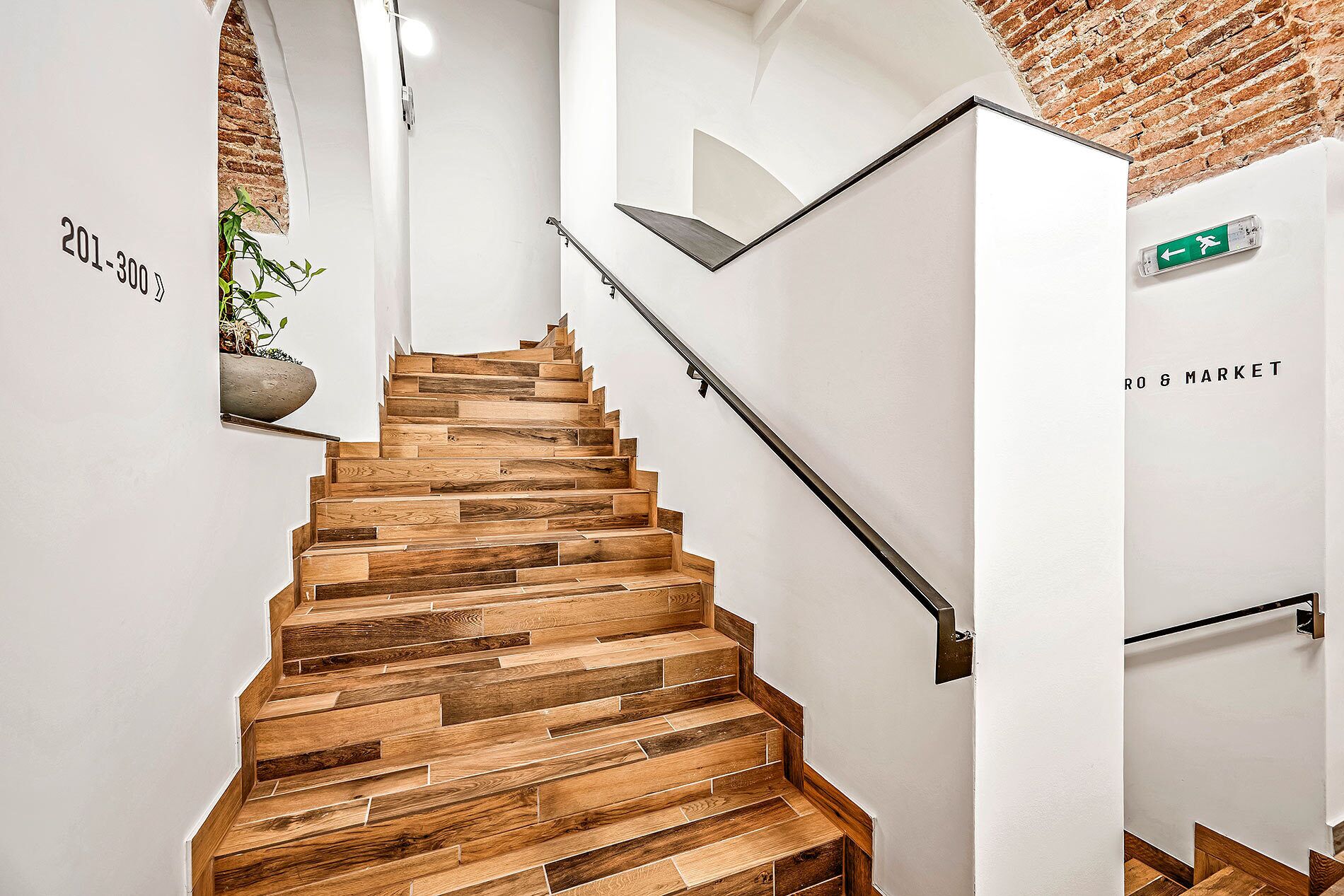 A staircase with wooden steps and a metal handrail, a brick wall, and a green emergency exit sign.