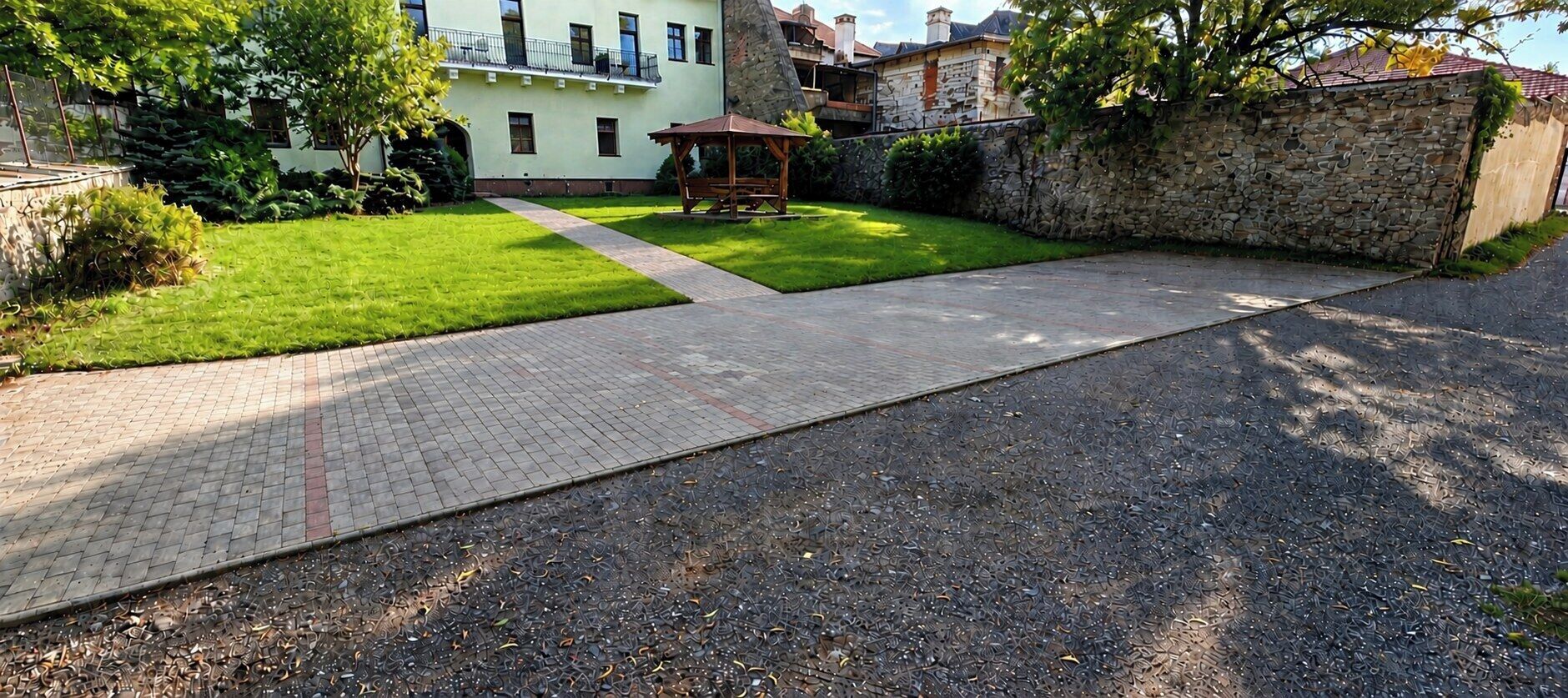 A paved pathway leading to a gazebo in a well-maintained garden area with a stone wall and a building in the background.