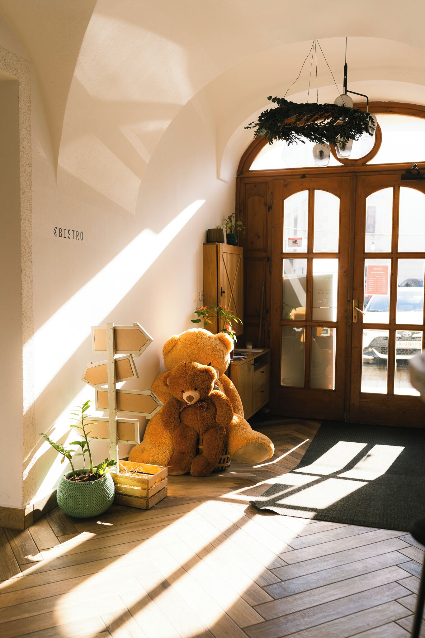 A room with a large teddy bear, a wooden cabinet, a potted plant, and a window with sunlight.