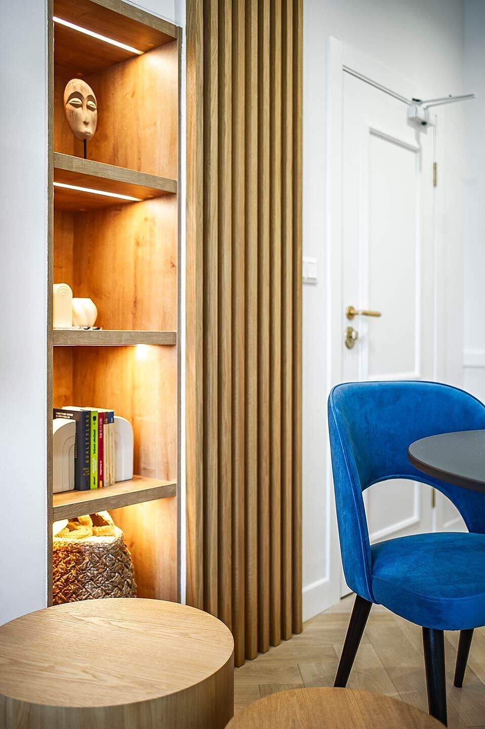 A room with a wooden bookshelf, a blue chair, a round wooden table, and a white door.