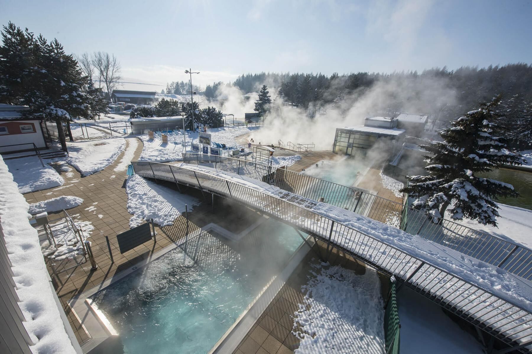Ein mit Schnee bedeckter Außenpoolbereich mit einer Brücke, umgeben von Bäumen und Gebäuden.