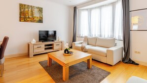 Un salon moderne avec une table basse en bois, un canapé beige, un téléviseur à écran plat posé sur un meuble en bois et un tableau représentant une forêt accroché au mur.