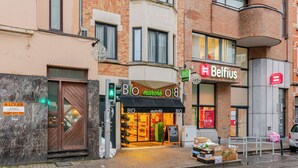 Une vue de rue avec un passage piéton, un panneau indiquant la limite de vitesse et des devantures de magasins, dont celles de « Belfius » et de « BO marché ».