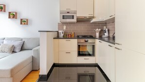 Une cuisine moderne équipée d’un four encastré, d’un four à micro-ondes et d’une cuisinière à gaz. Le salon adjacent est doté d’un canapé gris agrémenté de coussins.
