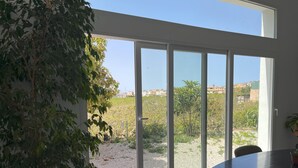A room with a large sliding glass door leading to a garden area. There is a round table with chairs, a potted plant, and a view of a residential area with houses and greenery.
