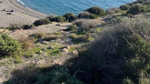 A coastal beach with a clear sky, rocky terrain, and a distant shoreline with buildings.
