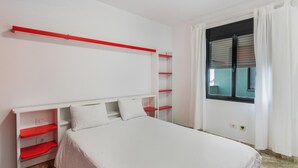 Un dormitorio con cama, paredes blancas, una ventana con cortinas y una estantería roja.