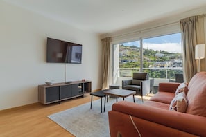 A modern living room with a flat-screen TV, a sofa, a coffee table, and a view of a cityscape through large windows.