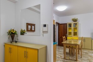 Ruokailutilassa on lasipöytä, puutuolit, keltainen kaappi, peili ja seinälle asennettu puhelin.