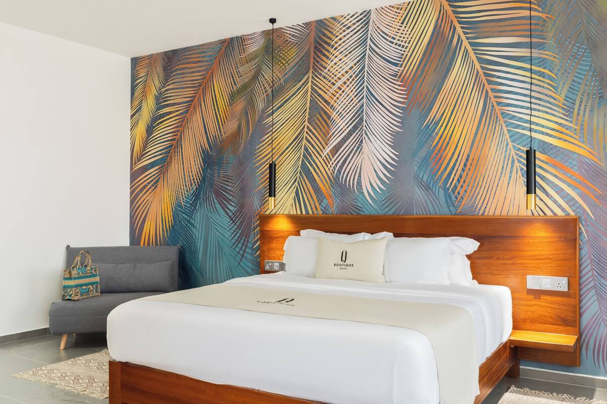 Une chambre d’hôtel moderne avec un grand lit, un canapé gris et une fresque murale aux motifs de feuilles de palmier aux couleurs vives.