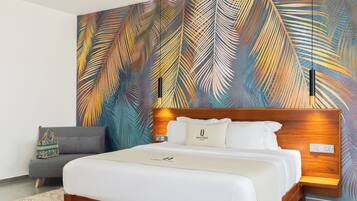 A modern hotel room with a large bed, a grey sofa, and a vibrant palm leaf mural on the wall.