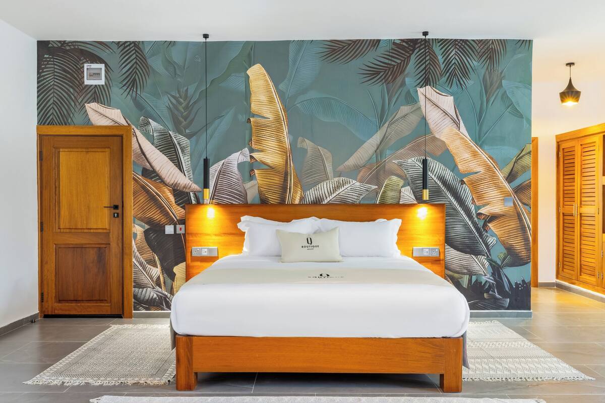 Une chambre avec un grand lit, une tête de lit en bois et une fresque murale représentant des feuilles tropicales.