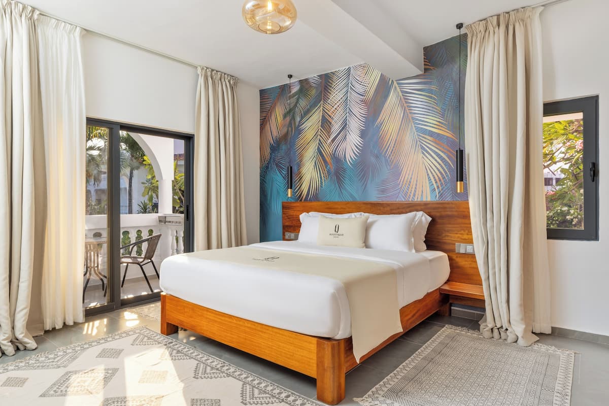 Une chambre d’hôtel moderne dotée d’un grand lit, d’une tête de lit en bois, d’une fresque représentant des feuilles tropicales et d’un balcon offrant une vue sur les palmiers.
