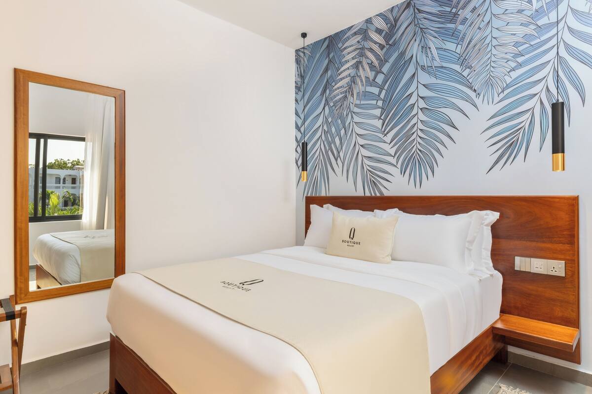 Une chambre d’hôtel avec un lit, une tête de lit en bois, un grand miroir et un mur orné d’un motif de feuilles.