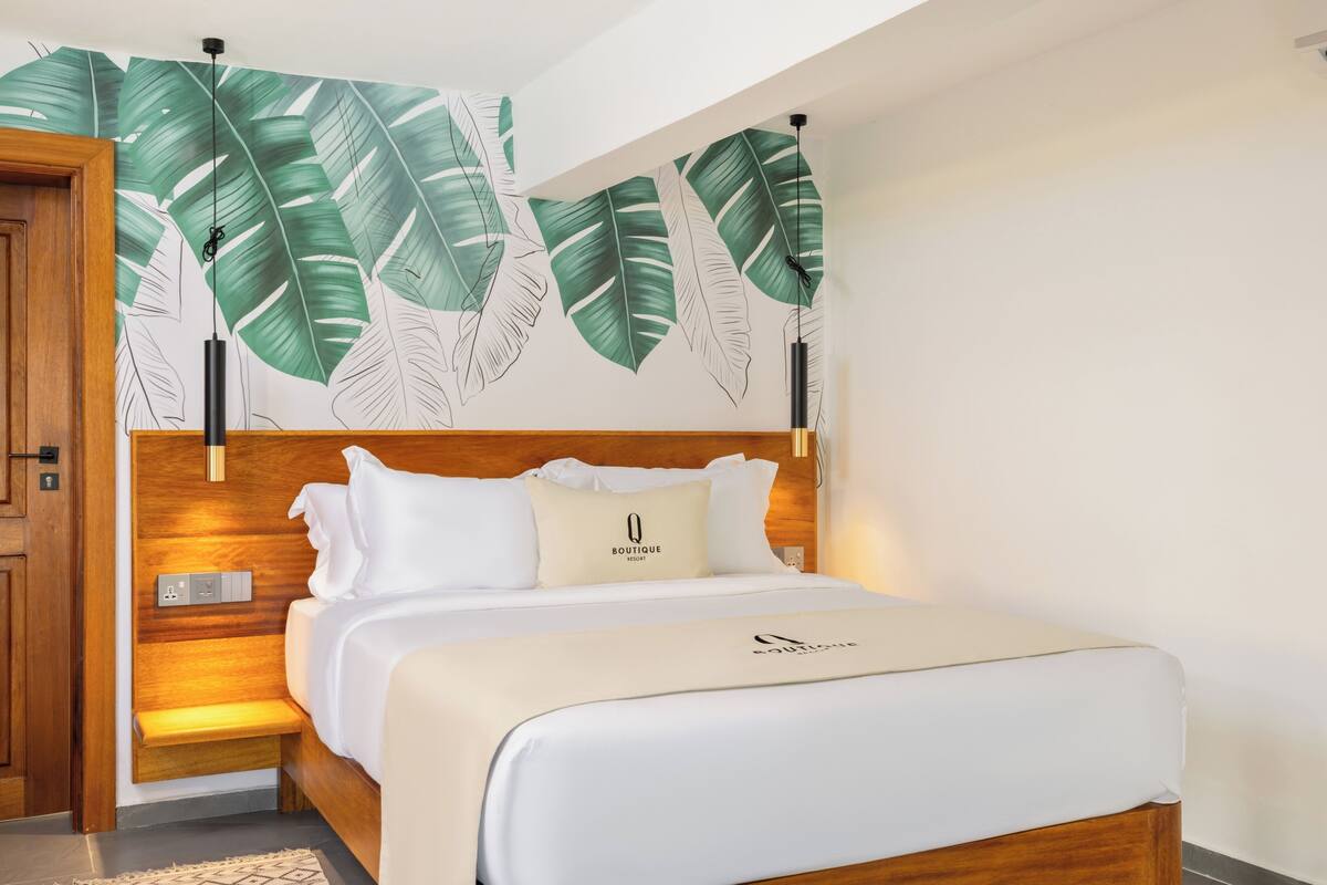 Une chambre d’hôtel avec une tête de lit en bois, un lit avec une literie blanche, une table de chevet et un mur orné d’un motif de feuilles.