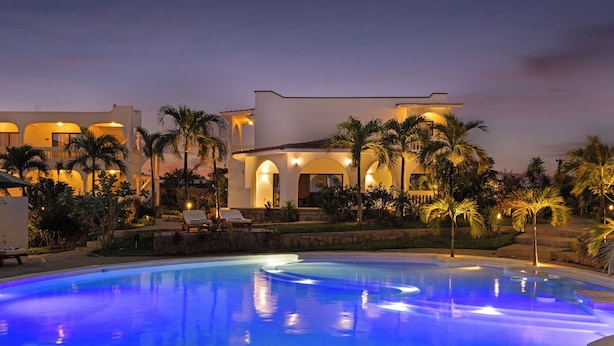 Una piscina con una zona junto a la piscina bien iluminada, palmeras y un edificio con varios balcones y ventanas de arco.