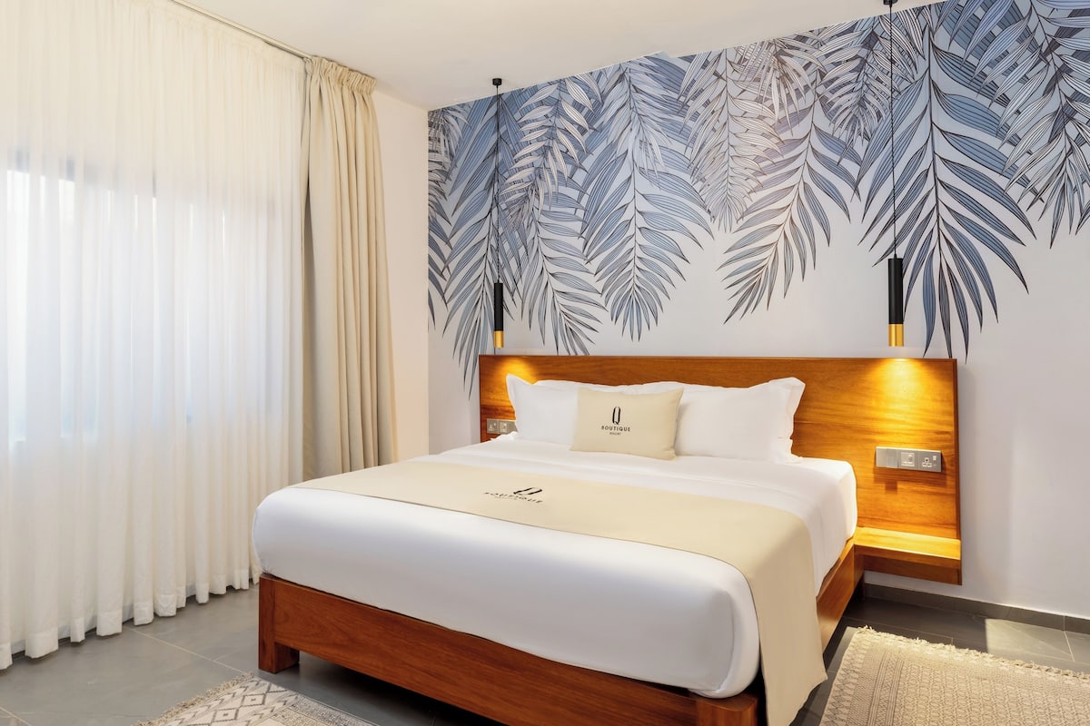 Une chambre d’hôtel moderne avec un grand lit, une tête de lit en bois, une fresque murale représentant des feuilles de palmier et une table de chevet avec une lampe.