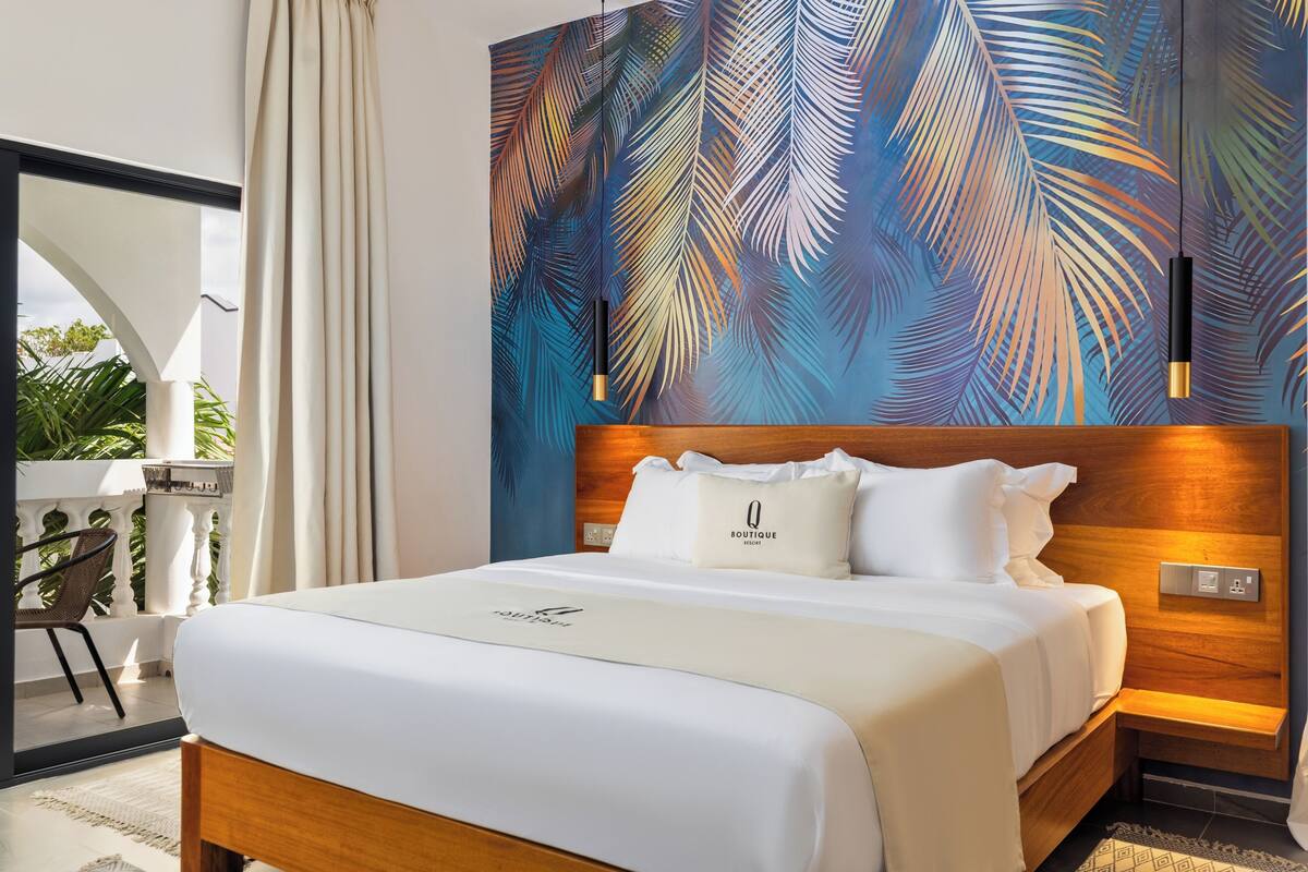 Une chambre d’hôtel avec un lit, une tête de lit en bois, un mur orné d’un motif de feuilles de palmier, un balcon avec une chaise et un luminaire suspendu.