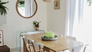 A dining area with a wooden table, white chairs, a woven pendant light, a round mirror, and a potted plant.
