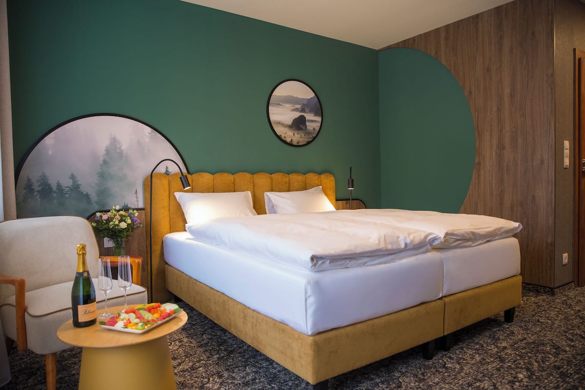 A hotel room with a bed, a round mirror, a wooden headboard, a small table with a bottle and glasses, and a plate of fruit.