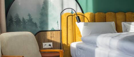Una habitación de hotel con una cama, una silla, una mesita con una taza y un platillo, y un mural en la pared.